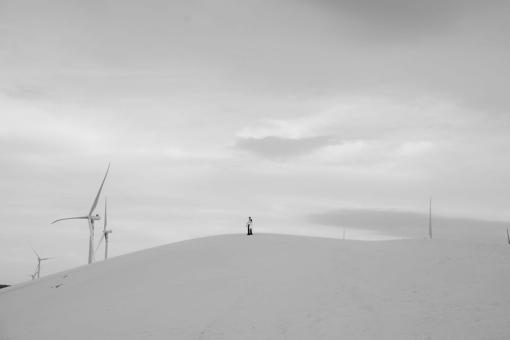 Black and white minimalist photo of a person standing alone on sand dunes with wind turbines.