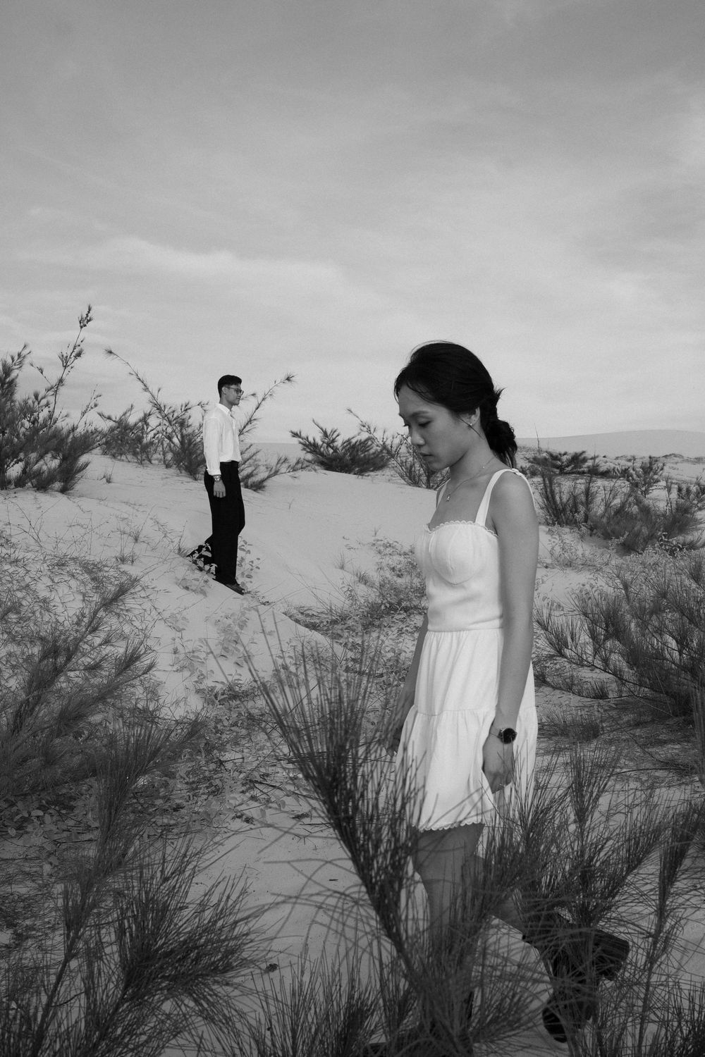 Black and white photo of a woman in white dress with man gesturing in sandy shrubland.