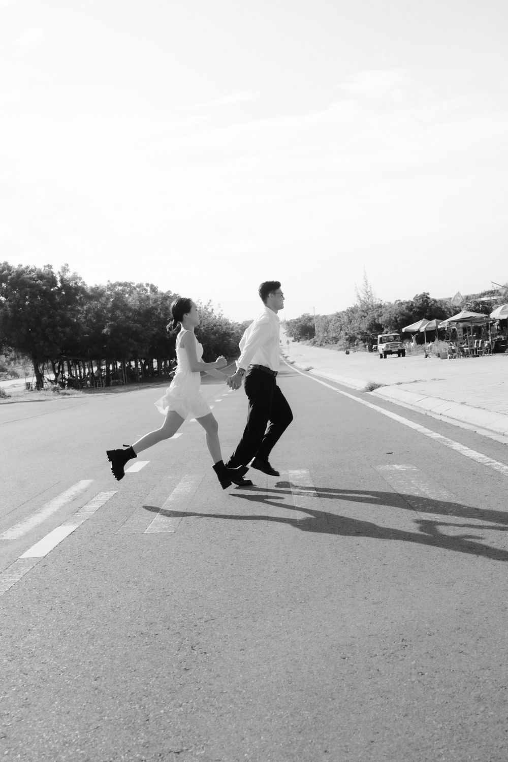 Black and white photo of a joyful couple running and holding hands across a sunny road.