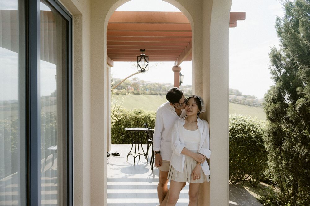 Romantic couple embracing on sunlit villa porch with garden and countryside views behind them.