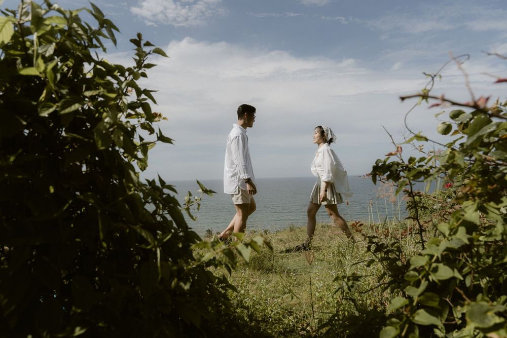 Couple in matching white outfits walking through lush coastal greenery with ocean view behind them.