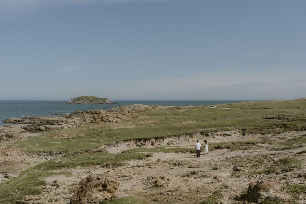 Couple in white attire walking across rocky coastal landscape with ocean and island in background.