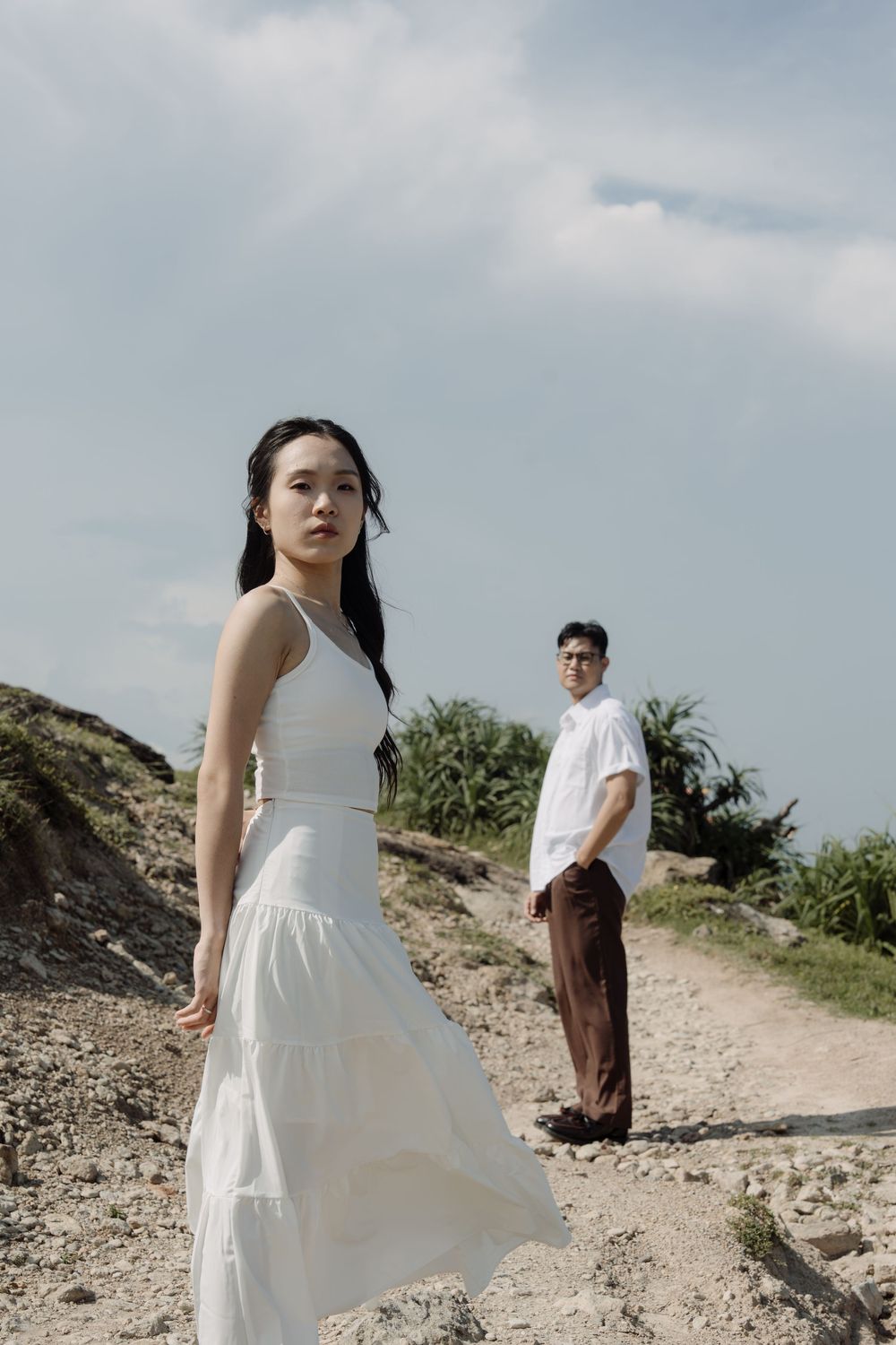 Woman in white dress stands in foreground as man in white shirt watches from rocky path.