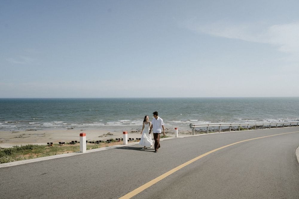 Couple holding hands walking along coastal road with vast ocean horizon stretching behind them.