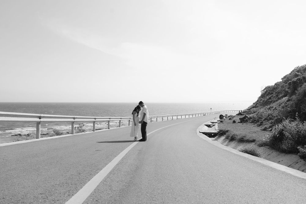 Black and white photo of couple kissing on coastal road with ocean and hillside views.