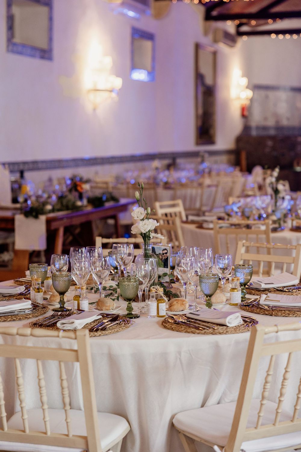 Elegant wedding reception tables set with white linens, glassware, and floral centerpieces in a warmly lit venue.