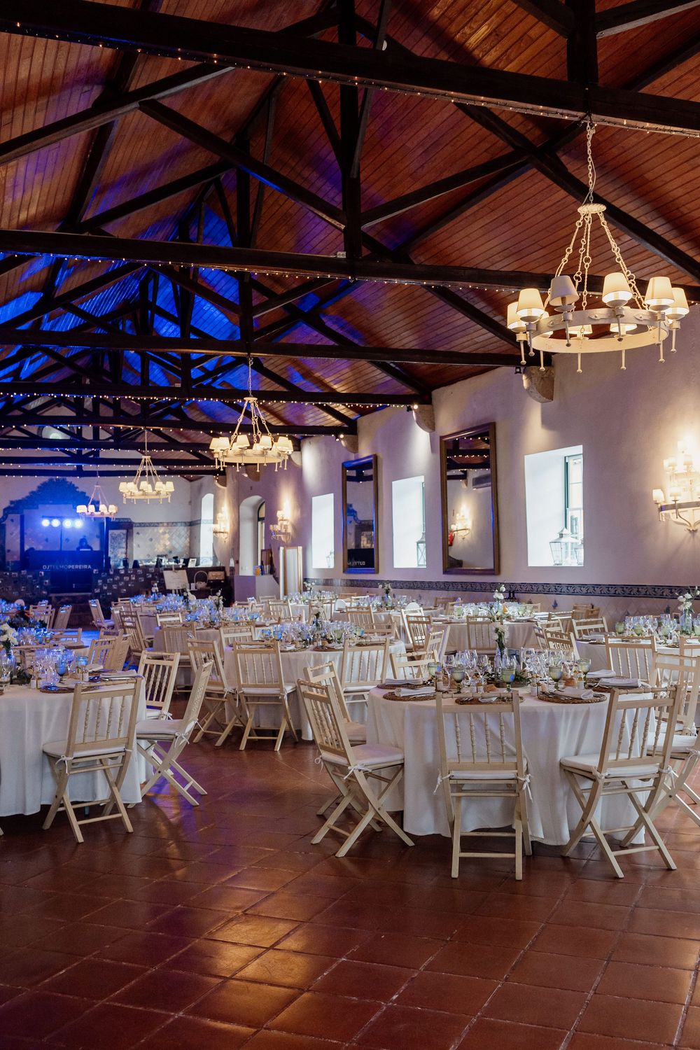 Elegant wedding reception hall with white chairs, round tables, chandeliers, and blue ambient lighting under a wooden vaulted ceiling.