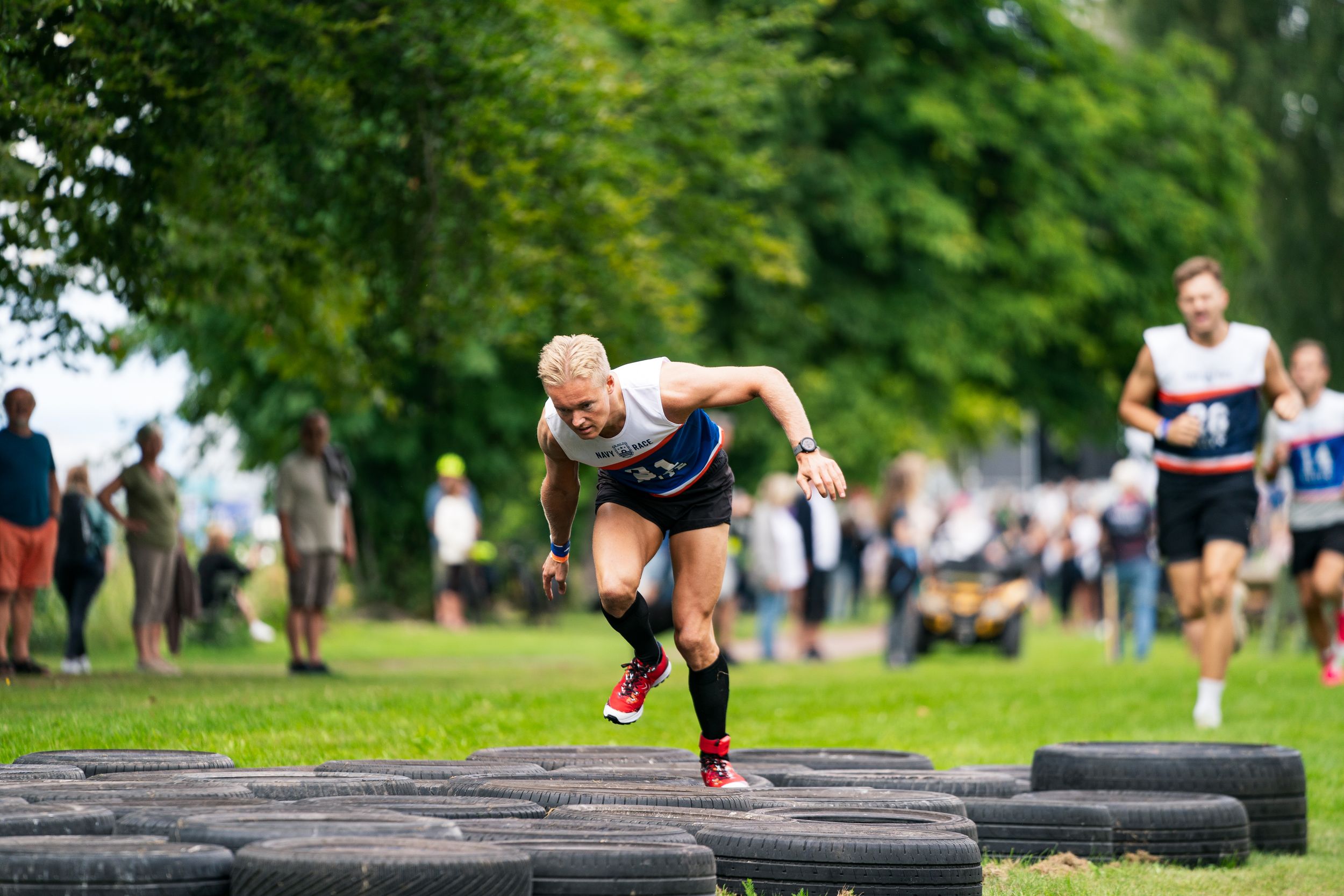Athlete sprinting over tires in an outdoor obstacle course race on a grassy field with spectators watching.