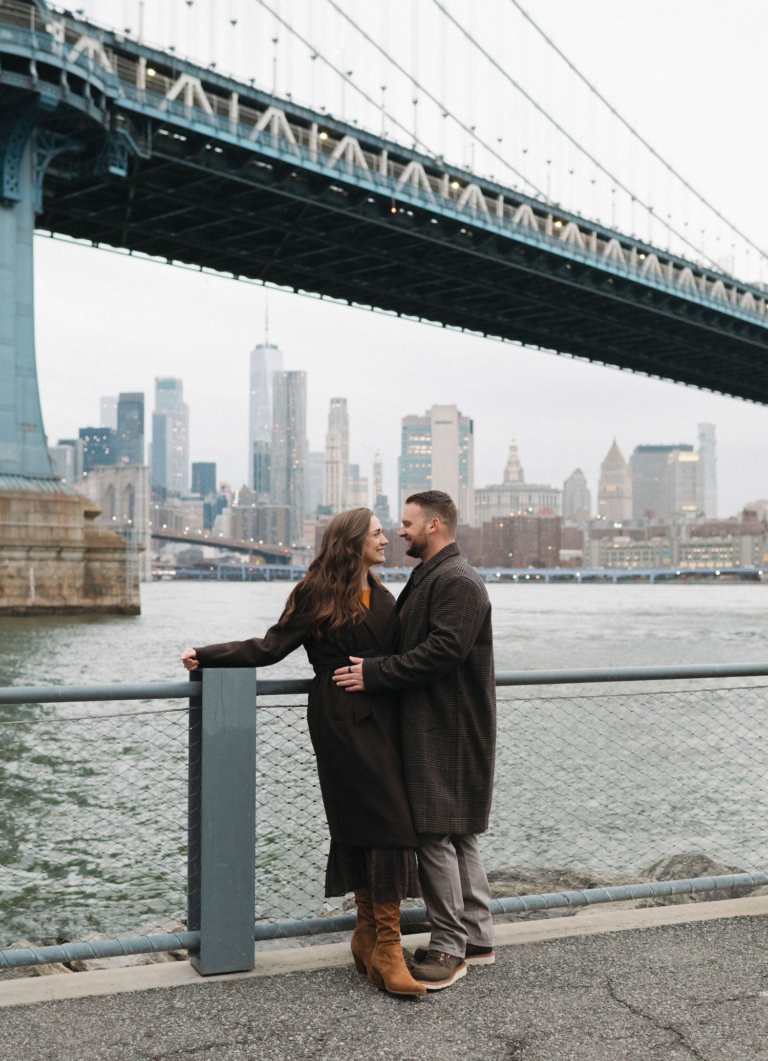 Couple embracing by the waterfront railing with the Manhattan Bridge and NYC skyline in the background.