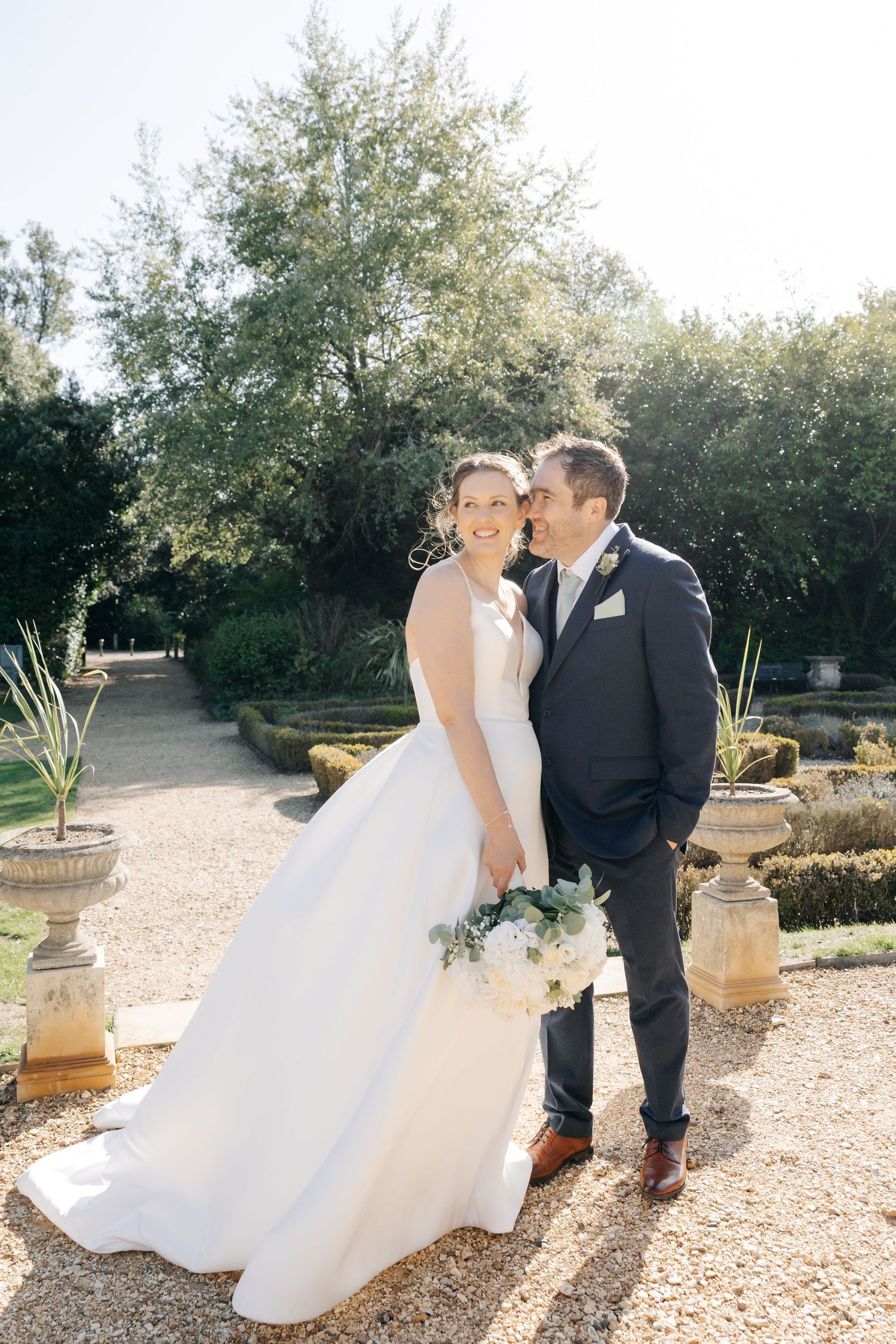 A couple in wedding attire pose together in a sunlit garden with stone urns and greenery in the background.