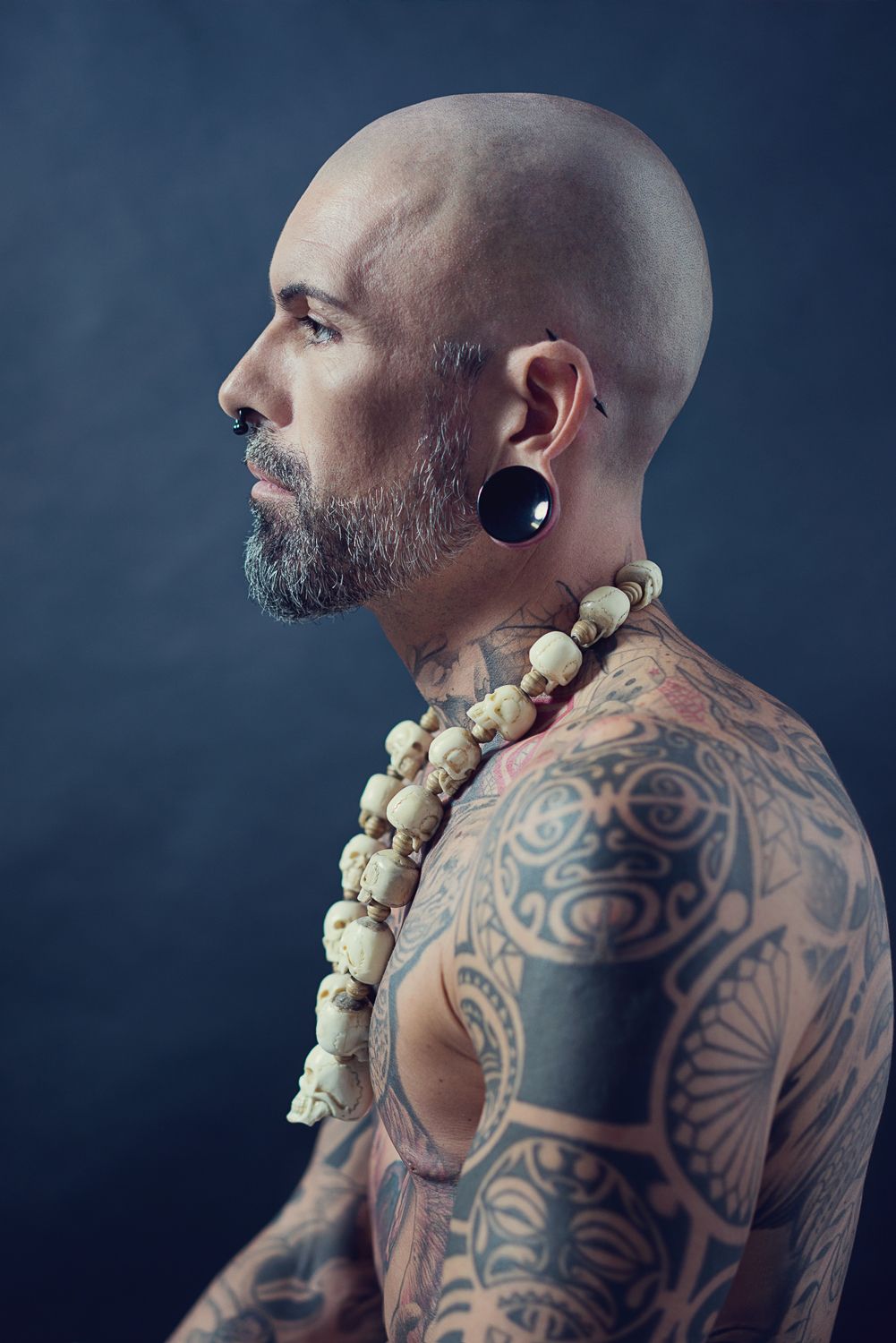 Profile portrait of bald tattooed man with beard, gauge earring, and skull necklace.