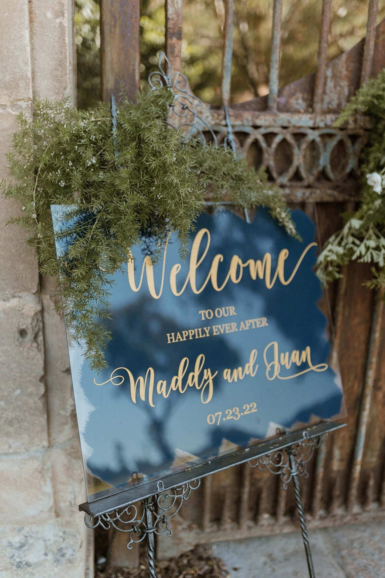 Gold calligraphy wedding welcome mirror sign for Maddy and Juan, dated 07.23.22, displayed on ornate iron easel.