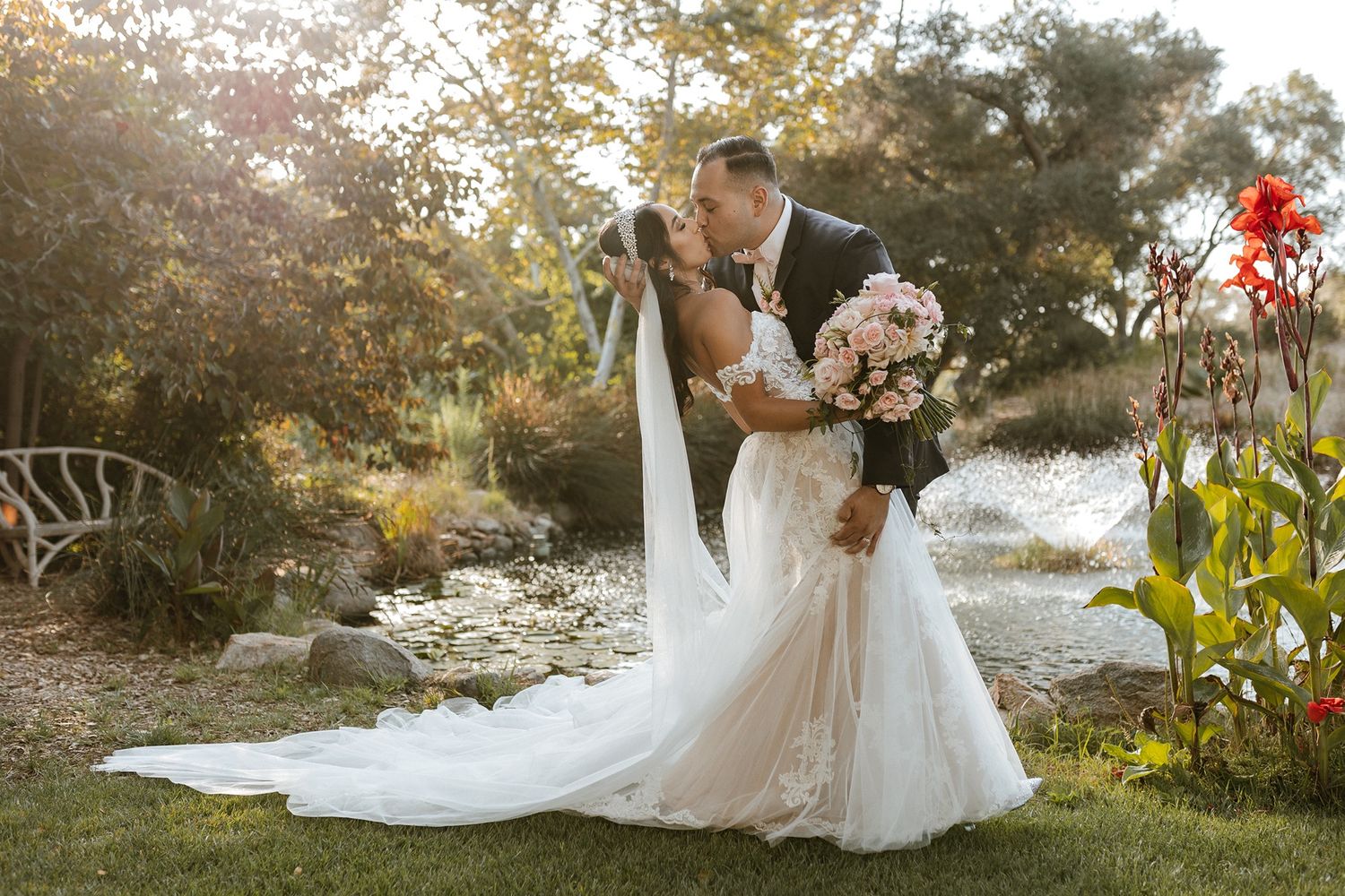 Bride and groom share a romantic kiss in a lush garden setting, with bride in elegant white gown and veil holding bouquet.