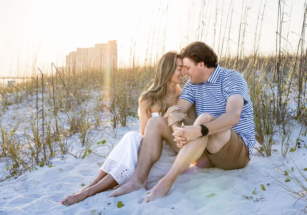 Couple sitting together on white sand beach dunes at sunset, sharing a romantic moment near coastal grass.