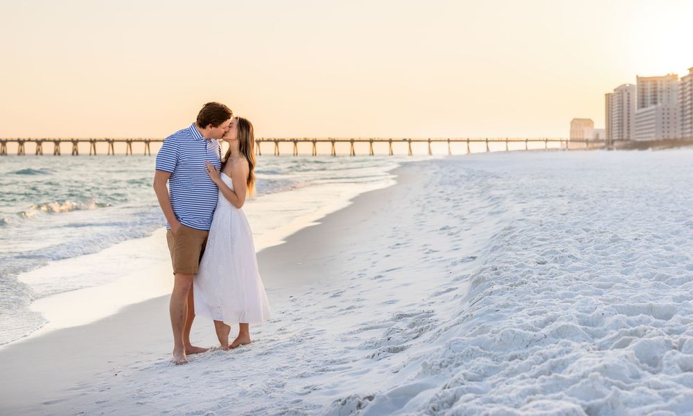 Romantic couple sharing a kiss on a white sandy beach at sunset with a pier in the background.
