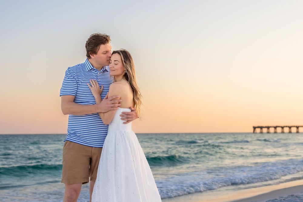 Couple embracing on a beach at sunset with ocean waves and pier in the background.