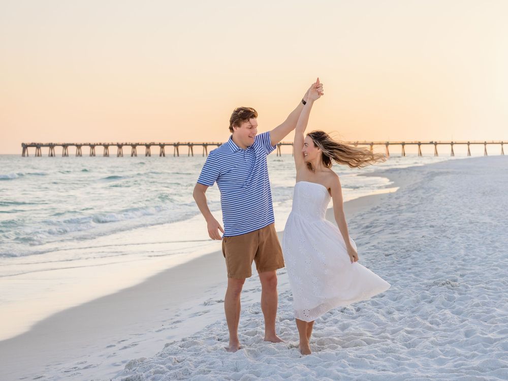 Couple dancing on a white sand beach at sunset with a pier in the background, woman in white dress.