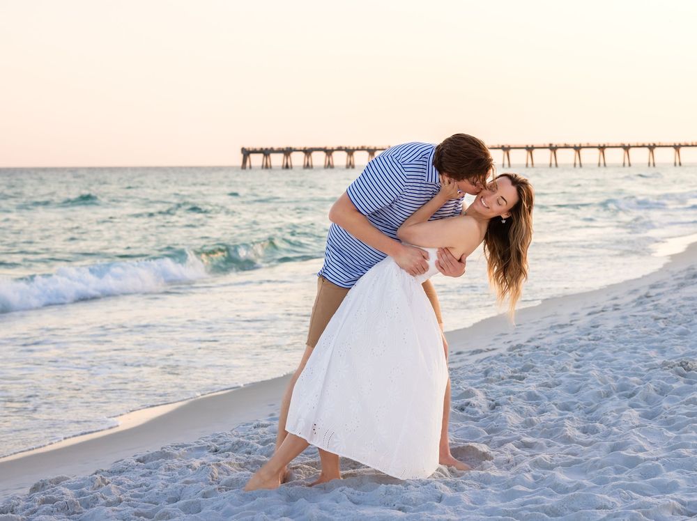 Romantic couple sharing a kiss on a sandy beach at sunset with a pier in the background.