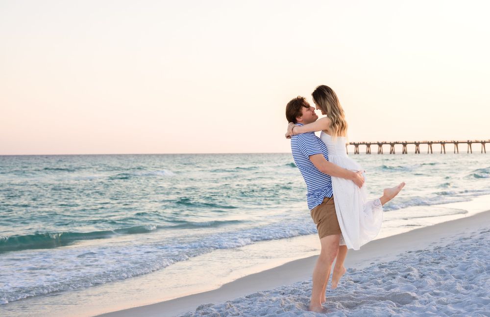 A couple embraces romantically on a sandy beach at sunset with a pier visible in the background.