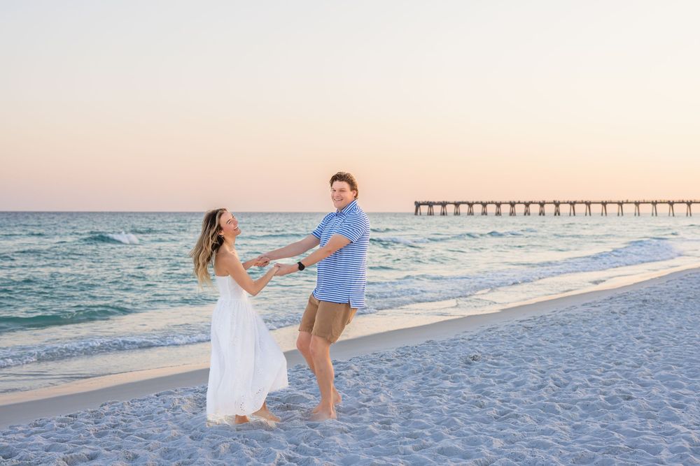 Couple dancing on a sandy beach at sunset near a pier, woman in white dress, man in blue shirt.