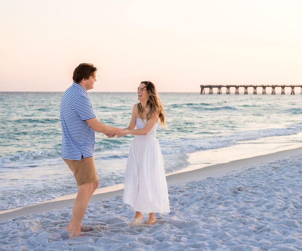 A couple holds hands and dances on a sandy beach at sunset with a pier in the background.