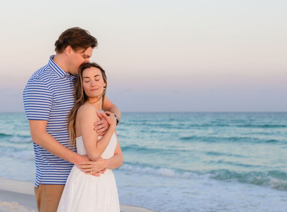 Couple embracing romantically on a beach at sunset, man in blue striped shirt hugging woman in white dress.