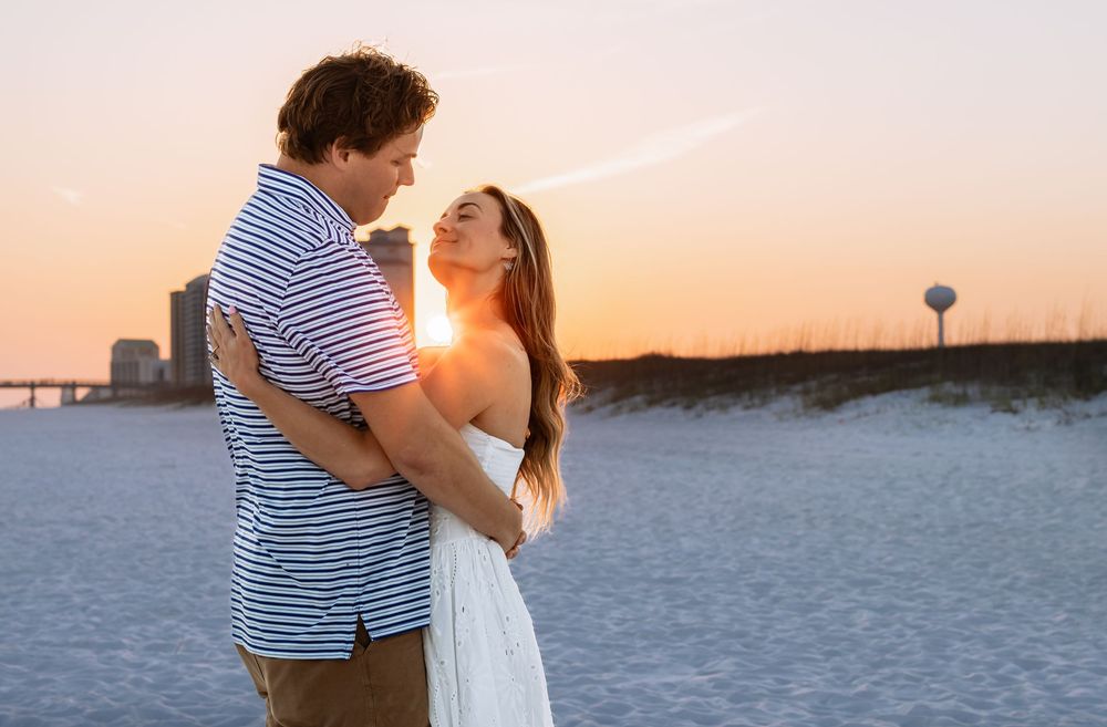Couple embracing on a sandy beach at golden sunset, woman in white dress, man in striped shirt, coastal backdrop.