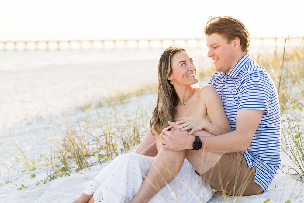 Couple sitting on beach sand dunes smiling, woman in white skirt, man in blue striped shirt, pier in background.