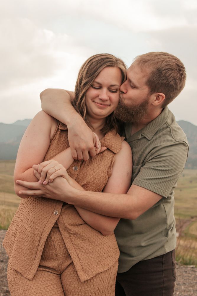 Bearded man in green shirt kissing smiling woman's cheek in a scenic mountain field | What to Wear for Family Photos