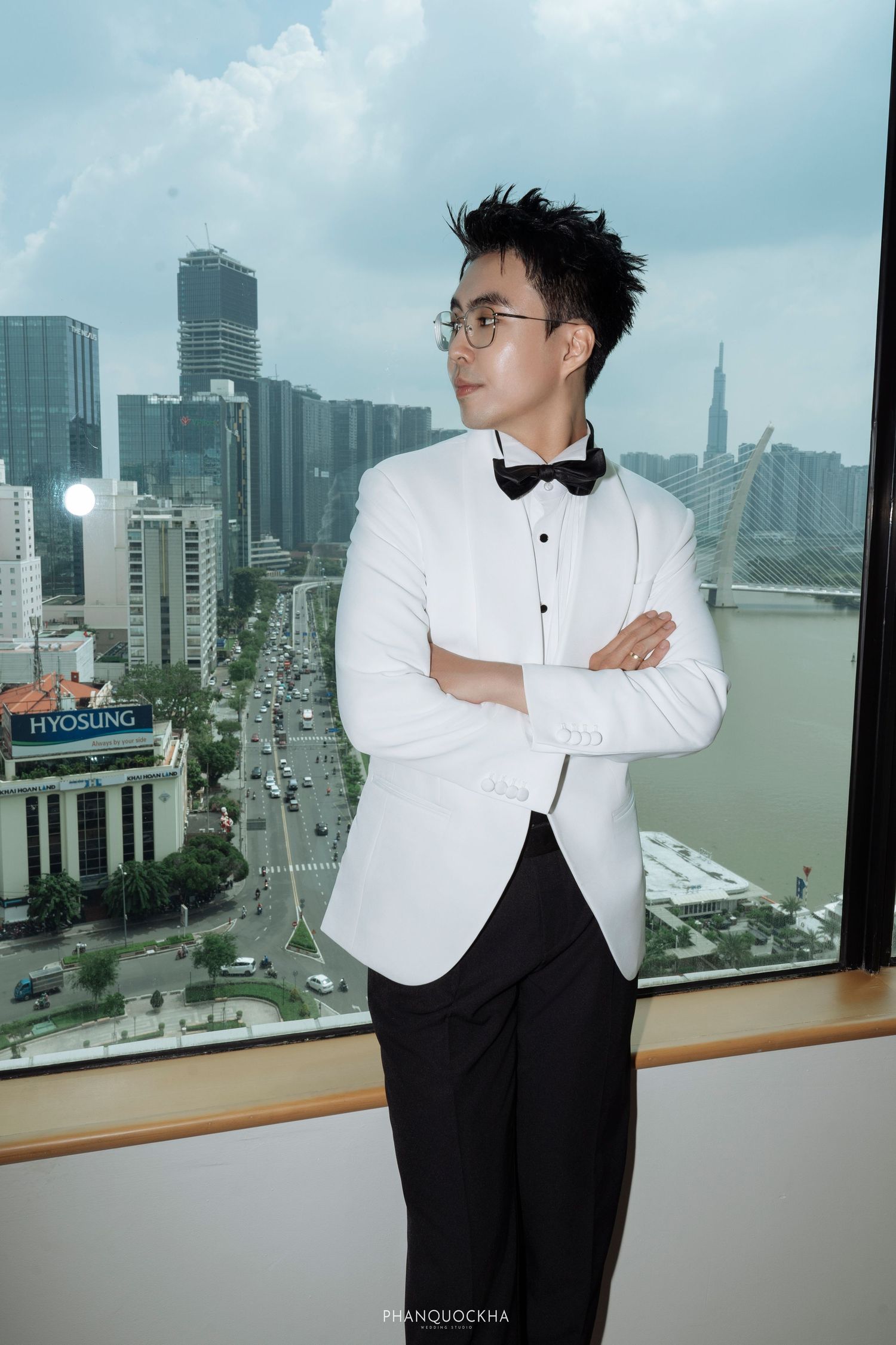 Groom in white tuxedo jacket and black bow tie stands with arms crossed by city skyline window.