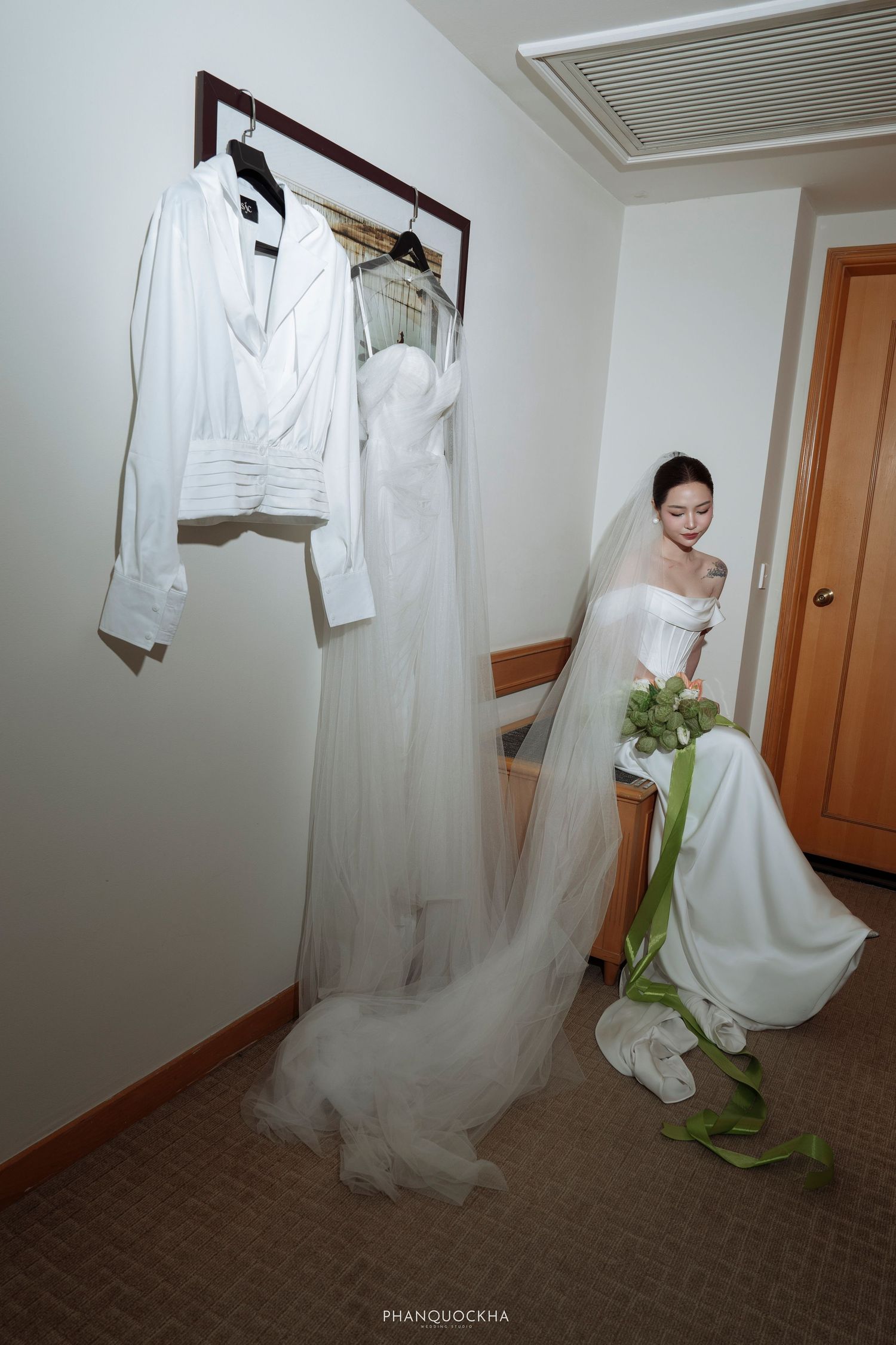 Bride in elegant white wedding gown holds green bouquet beside hanging bridal attire in hotel room.