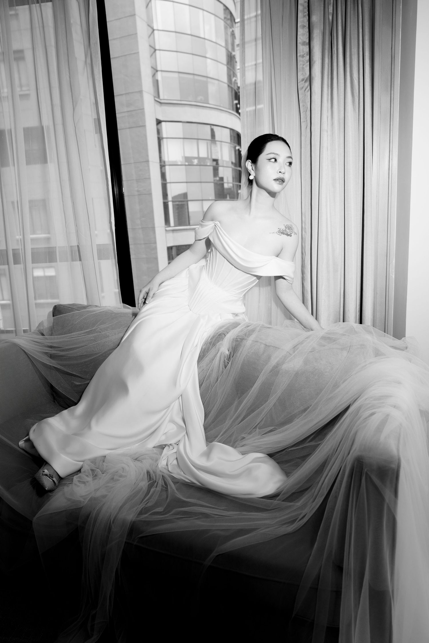 Black and white portrait of bride in off-shoulder wedding gown with flowing tulle by window.