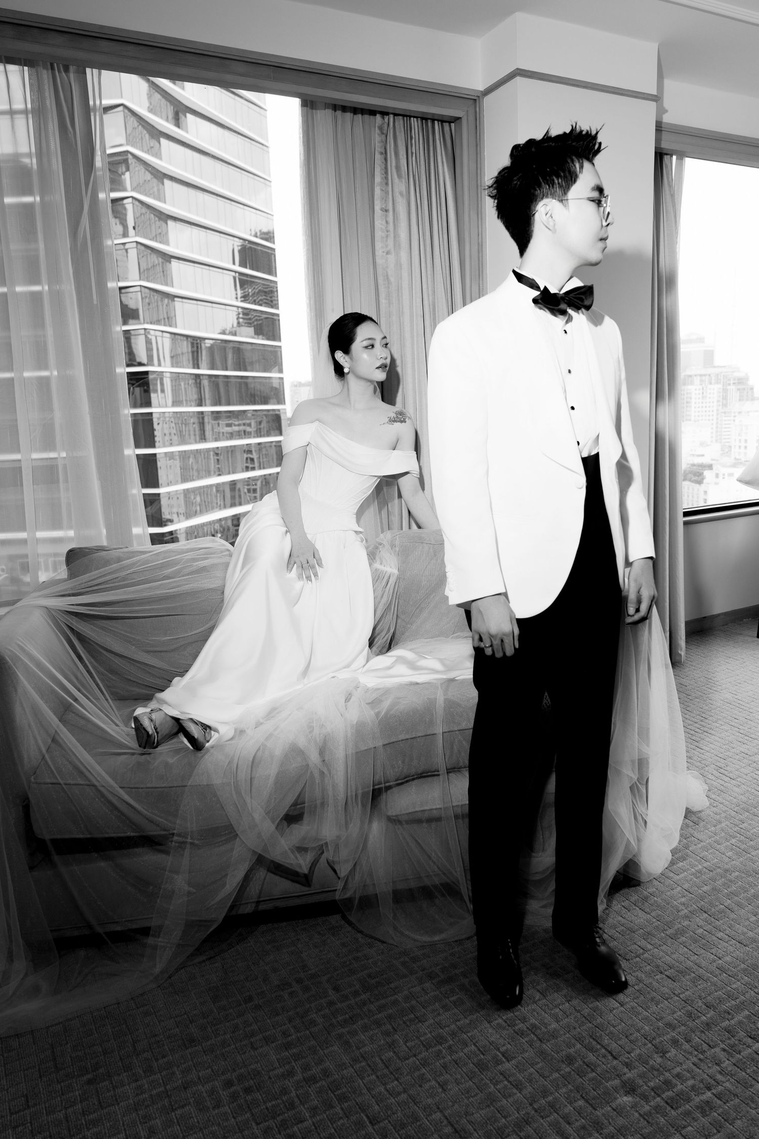 Black and white wedding photo of bride and groom posing separately in a hotel room with tulle veil.