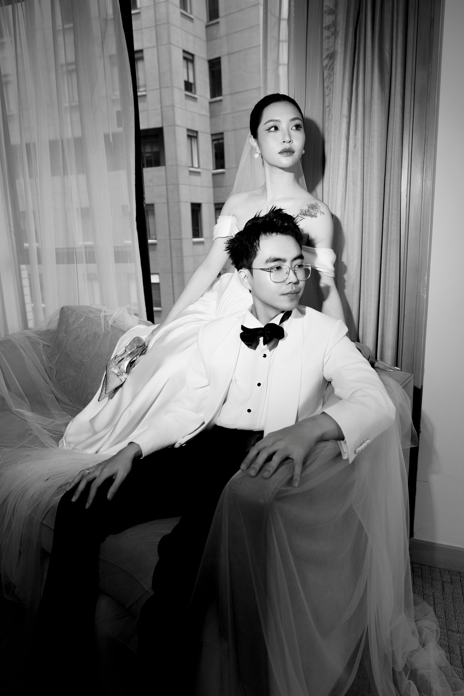Elegant black and white wedding portrait of couple posing together by curtained window on a sofa.