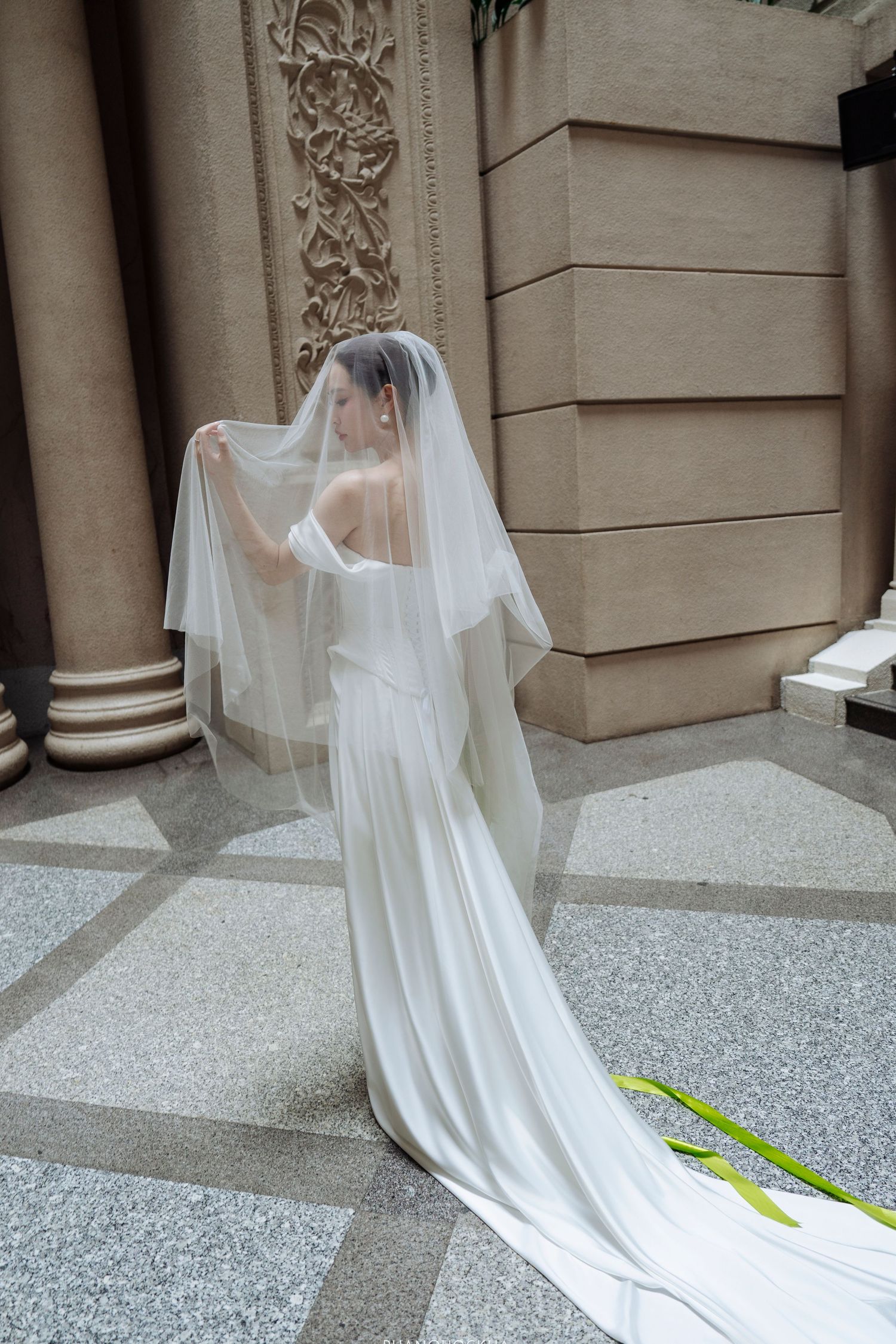 Bride in white off-shoulder gown lifts veil while standing before ornate stone columns outdoors.