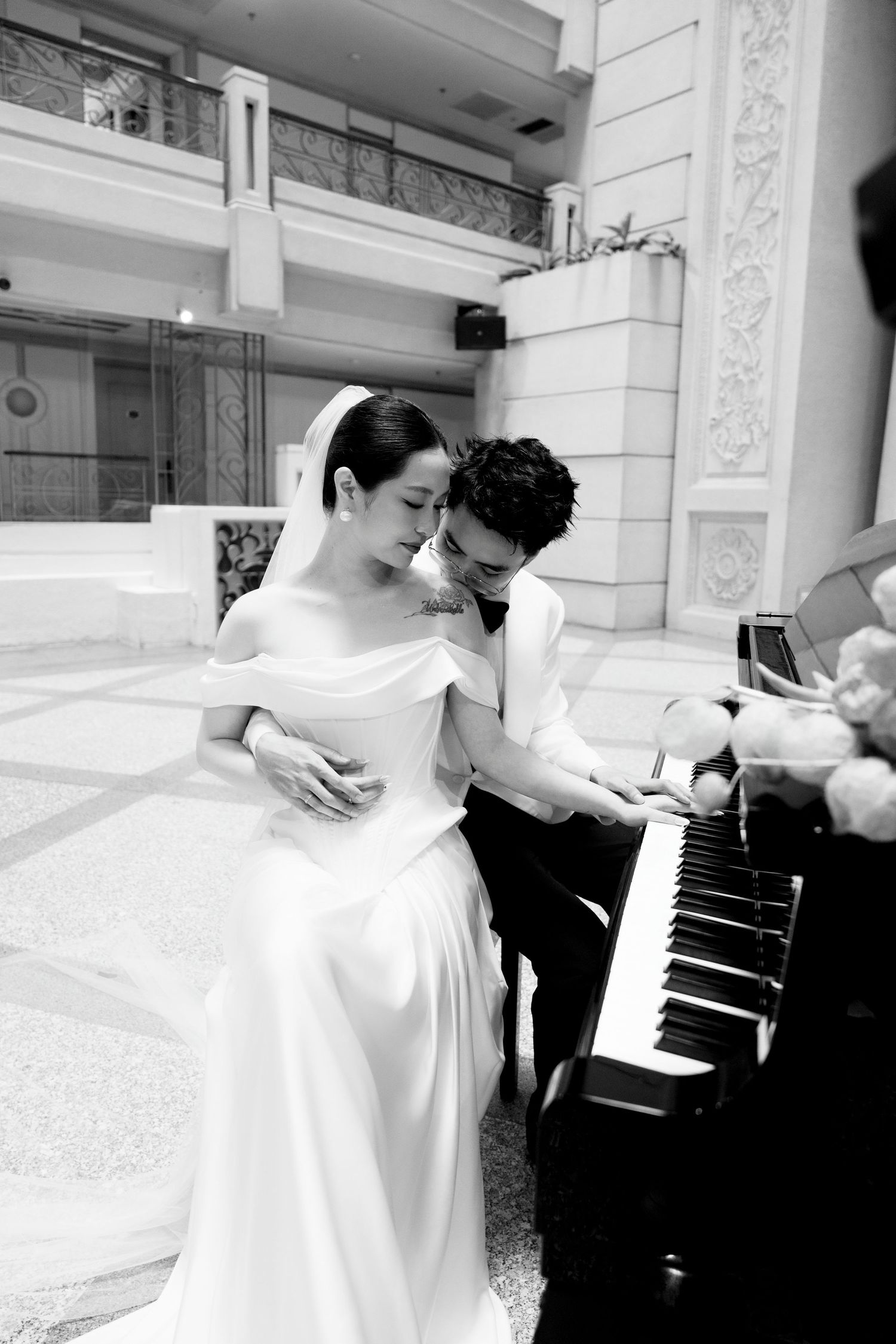 Romantic black and white wedding portrait of couple embracing at an upright piano in a grand hall.