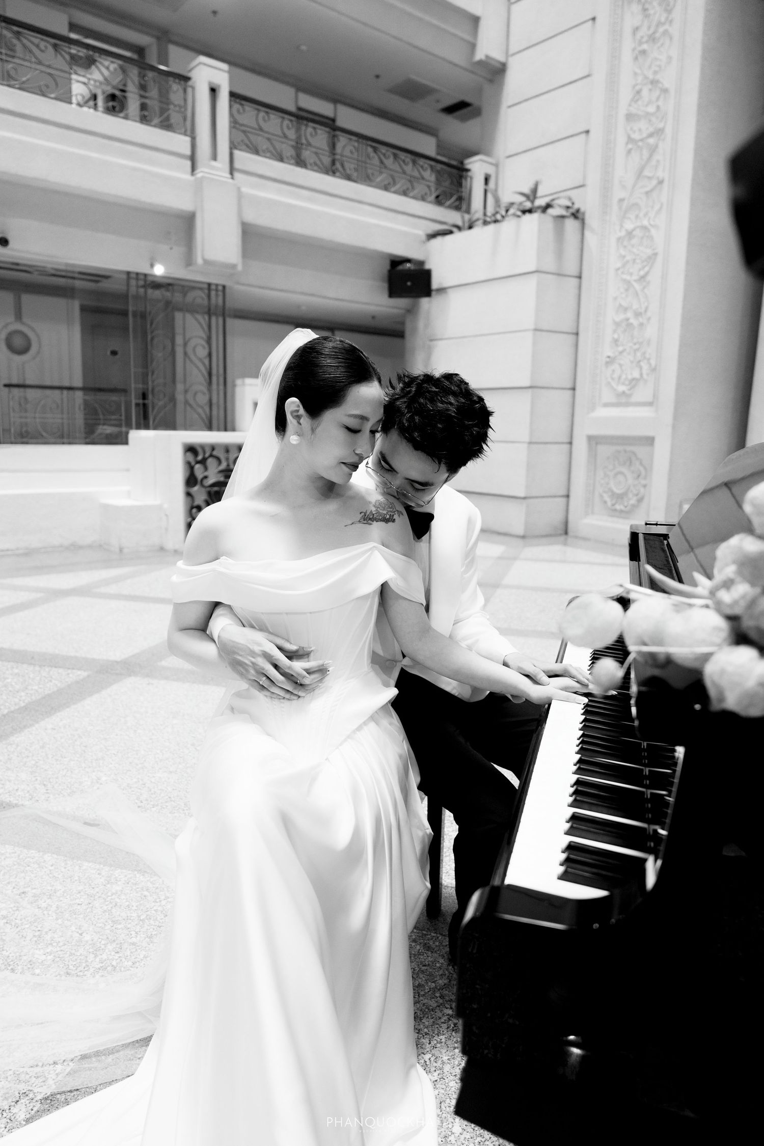 Black and white wedding photo of couple sharing romantic moment at a grand piano in elegant venue.