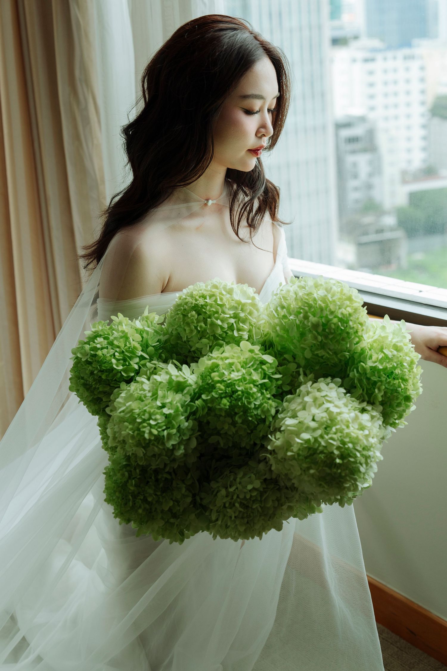 Beautiful bride in white gown holds oversized green hydrangea bouquet beside a sunlit window.