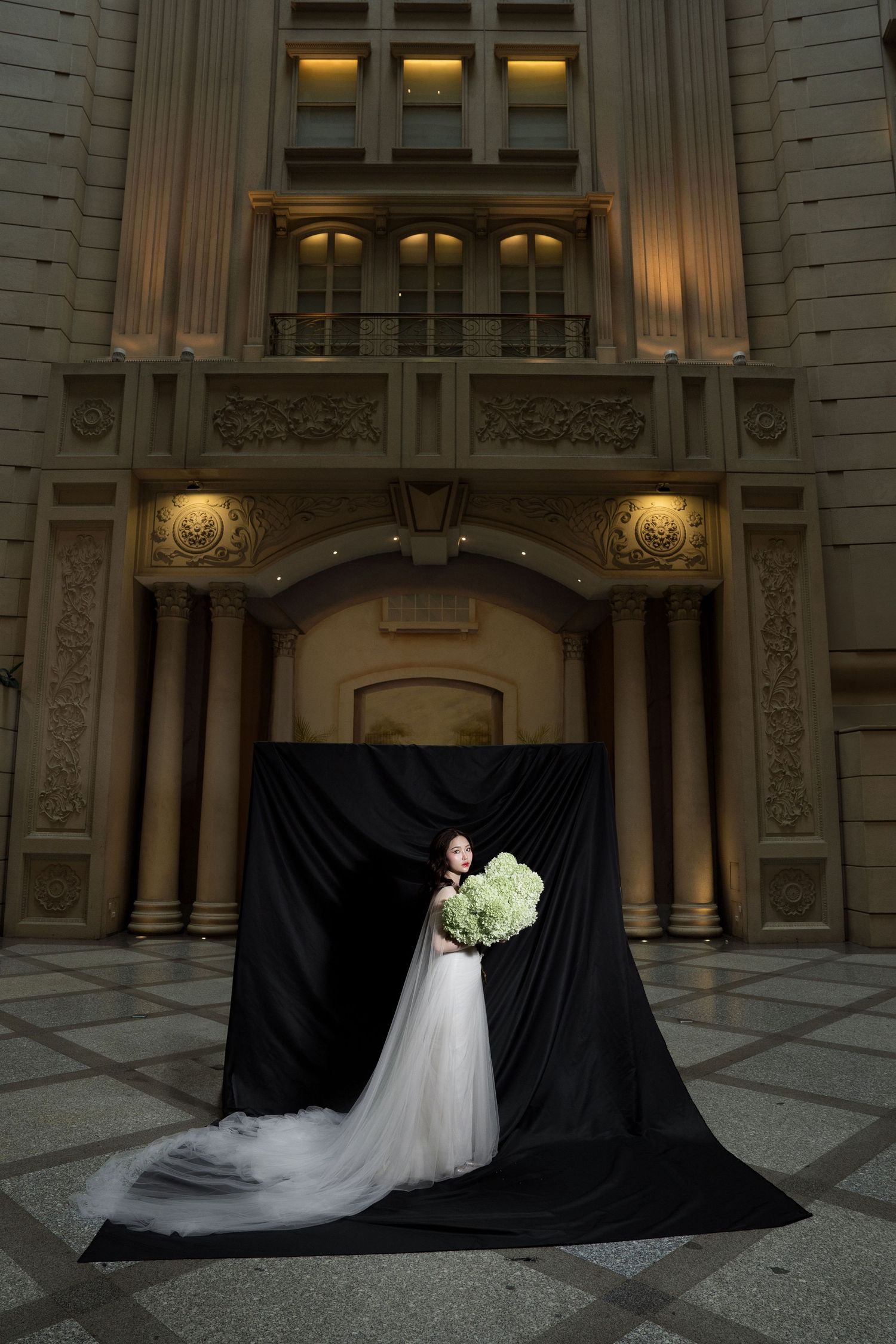 Bride in white gown holds green bouquet before dramatic black drape in ornate architectural setting.
