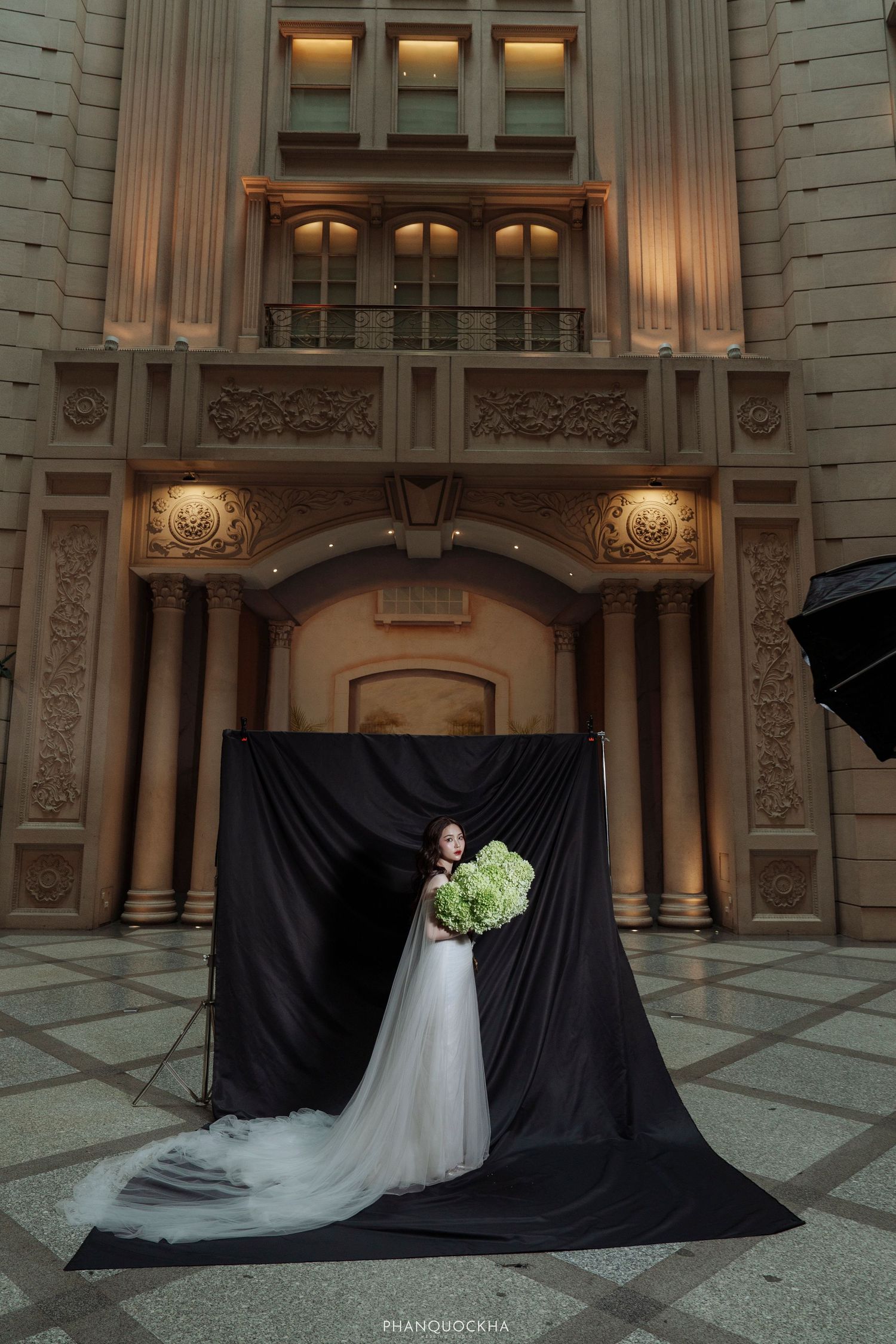Bride in white wedding gown holding green bouquet poses before black drape backdrop at ornate building.