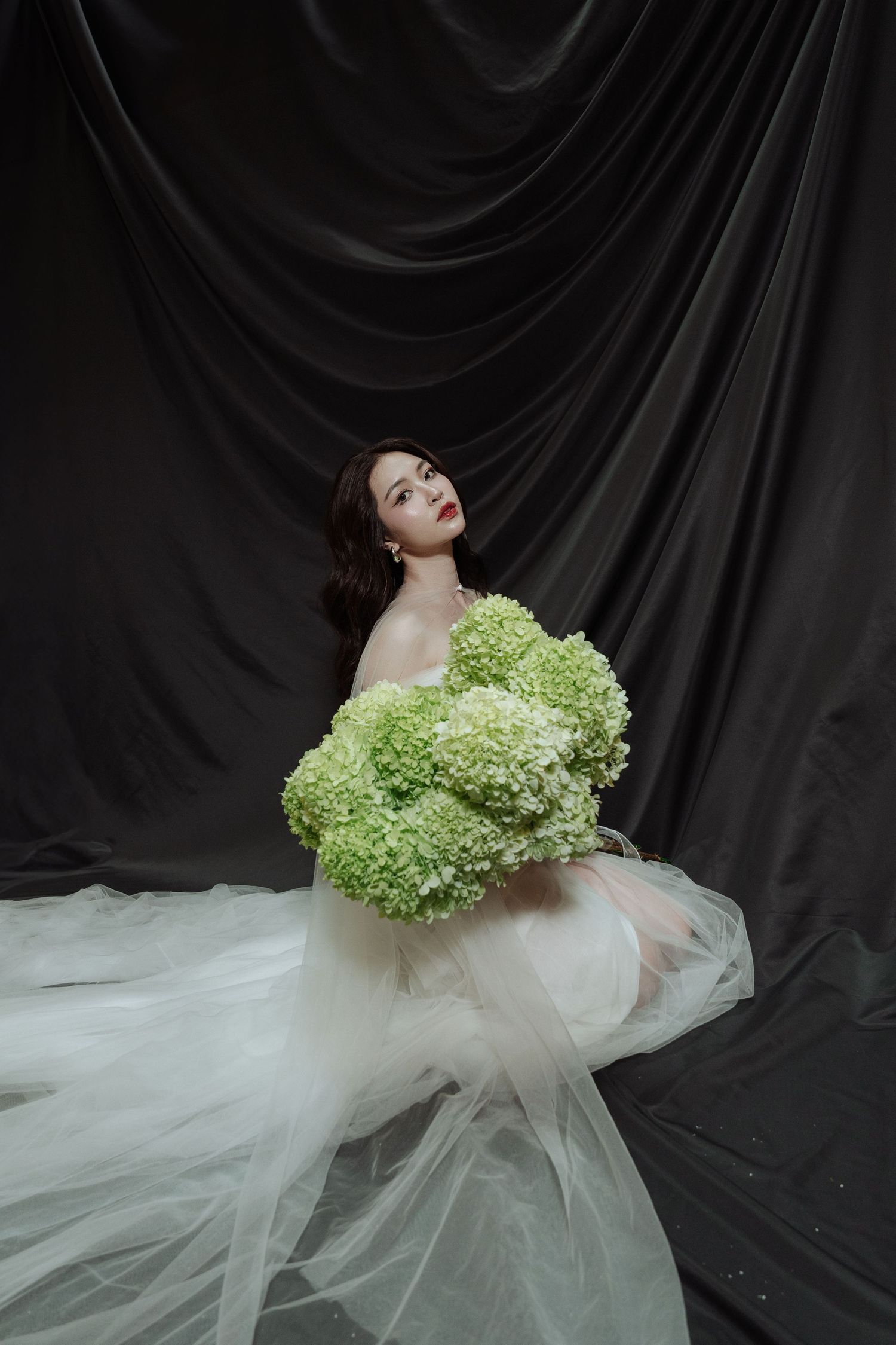 Bride in sheer white gown poses with large green hydrangea bouquet against flowing black fabric backdrop.