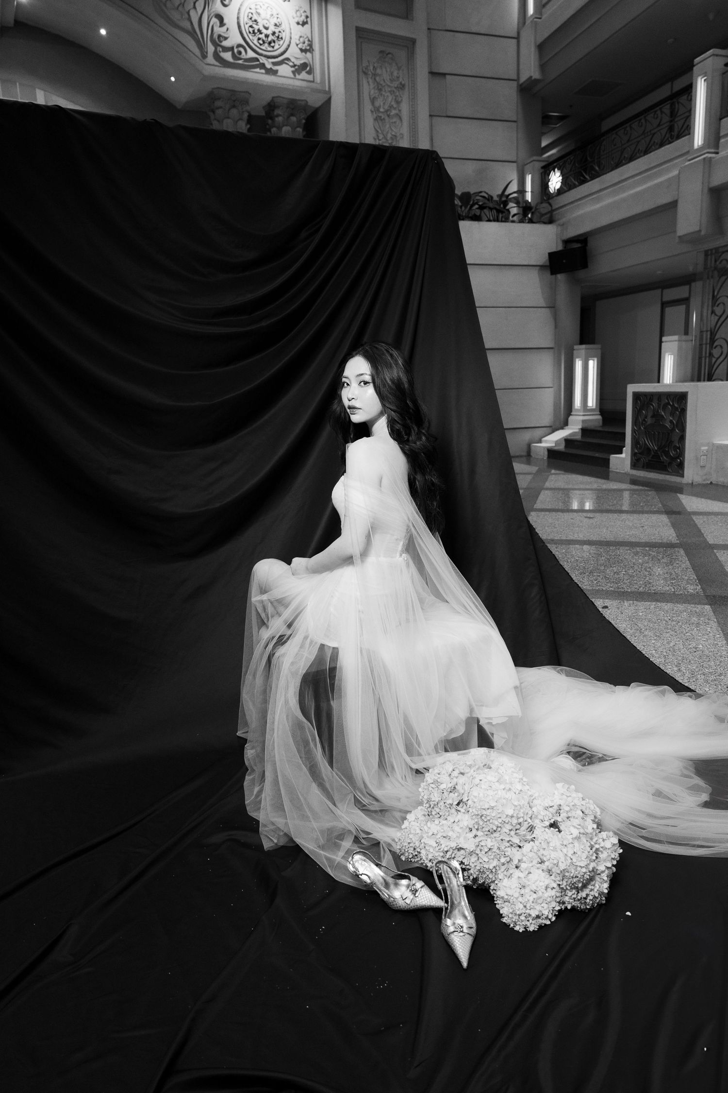 Black and white photo of bride in tulle gown seated on black drape with bouquet and heels.