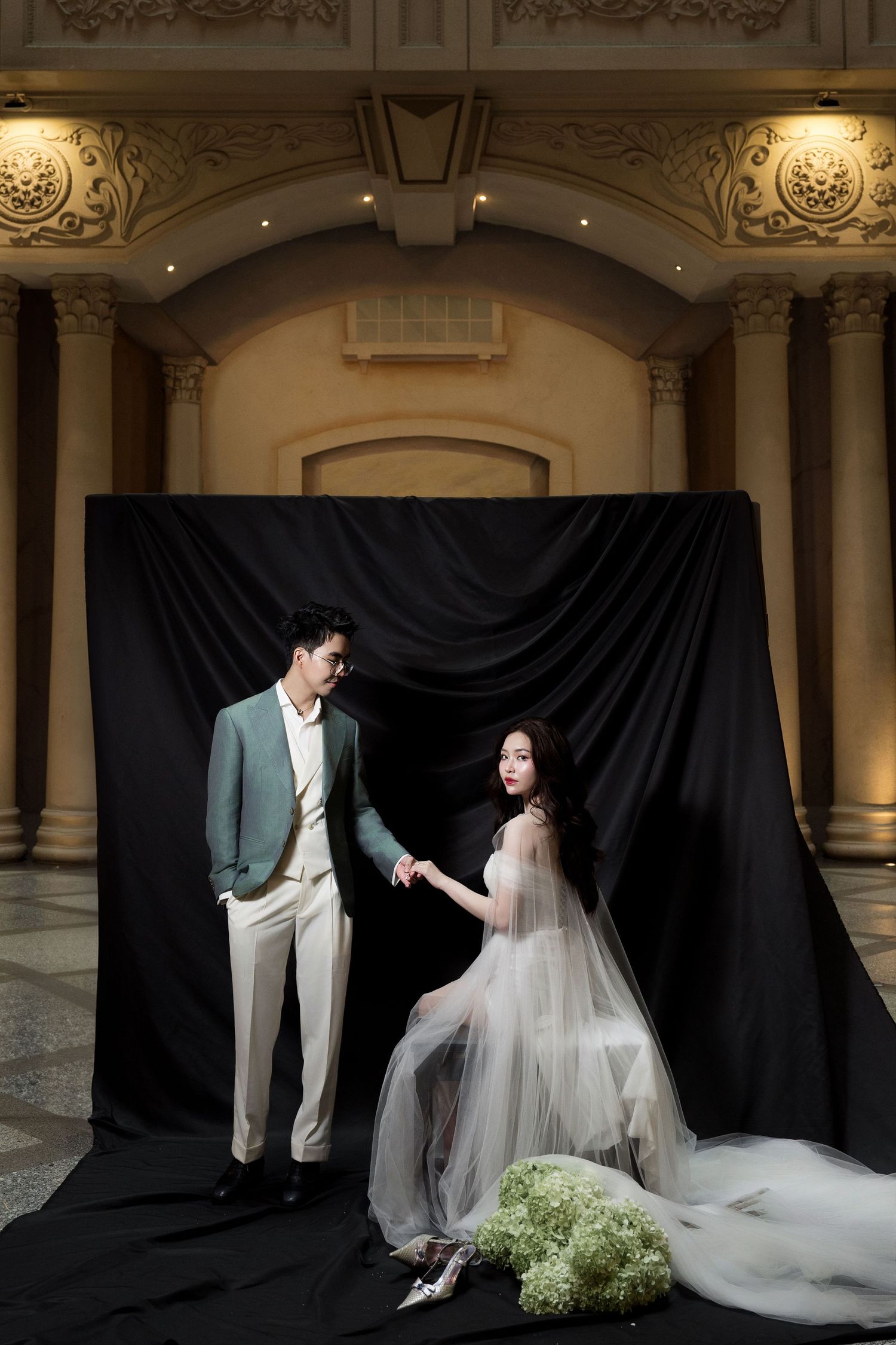Couple in elegant attire pose before black drape backdrop in a warmly lit ornate venue interior.