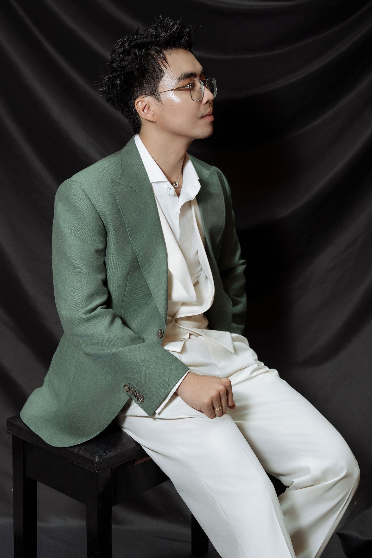 Young Asian man in green blazer and white pants sits on stool against black draped background.