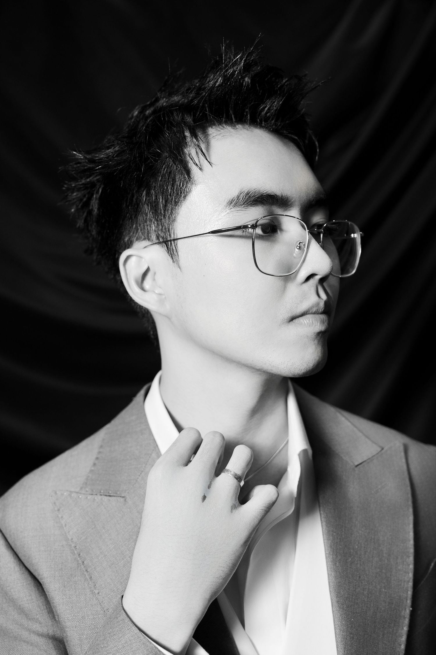 Black and white portrait of stylish young man in blazer and glasses adjusting collar against dark backdrop.