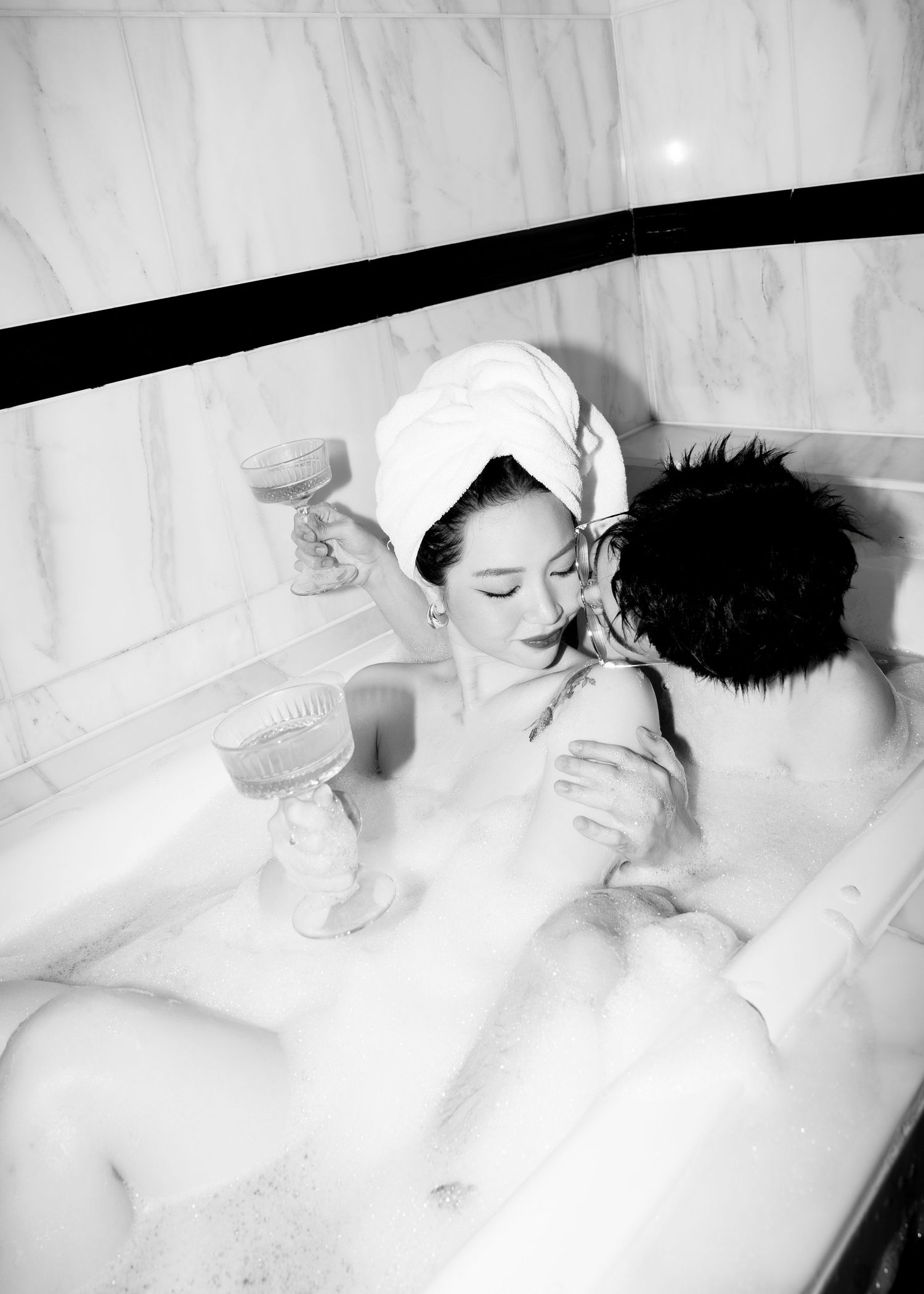 Black and white photo of couple sharing a romantic moment in a marble bathtub with champagne glasses.