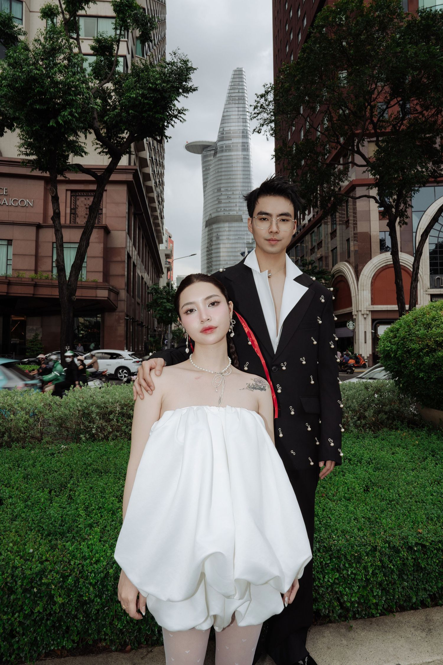 Couple in contrasting white and black outfits pose outdoors with Bitexco Financial Tower in background.