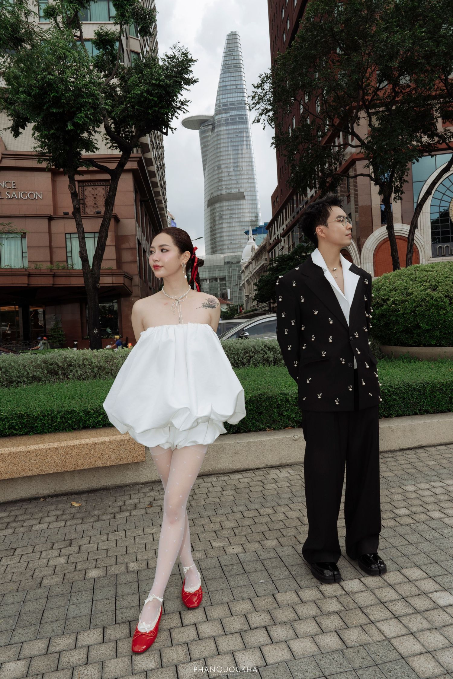 Couple poses in urban setting with woman in white bubble dress and red heels, man in black suit.