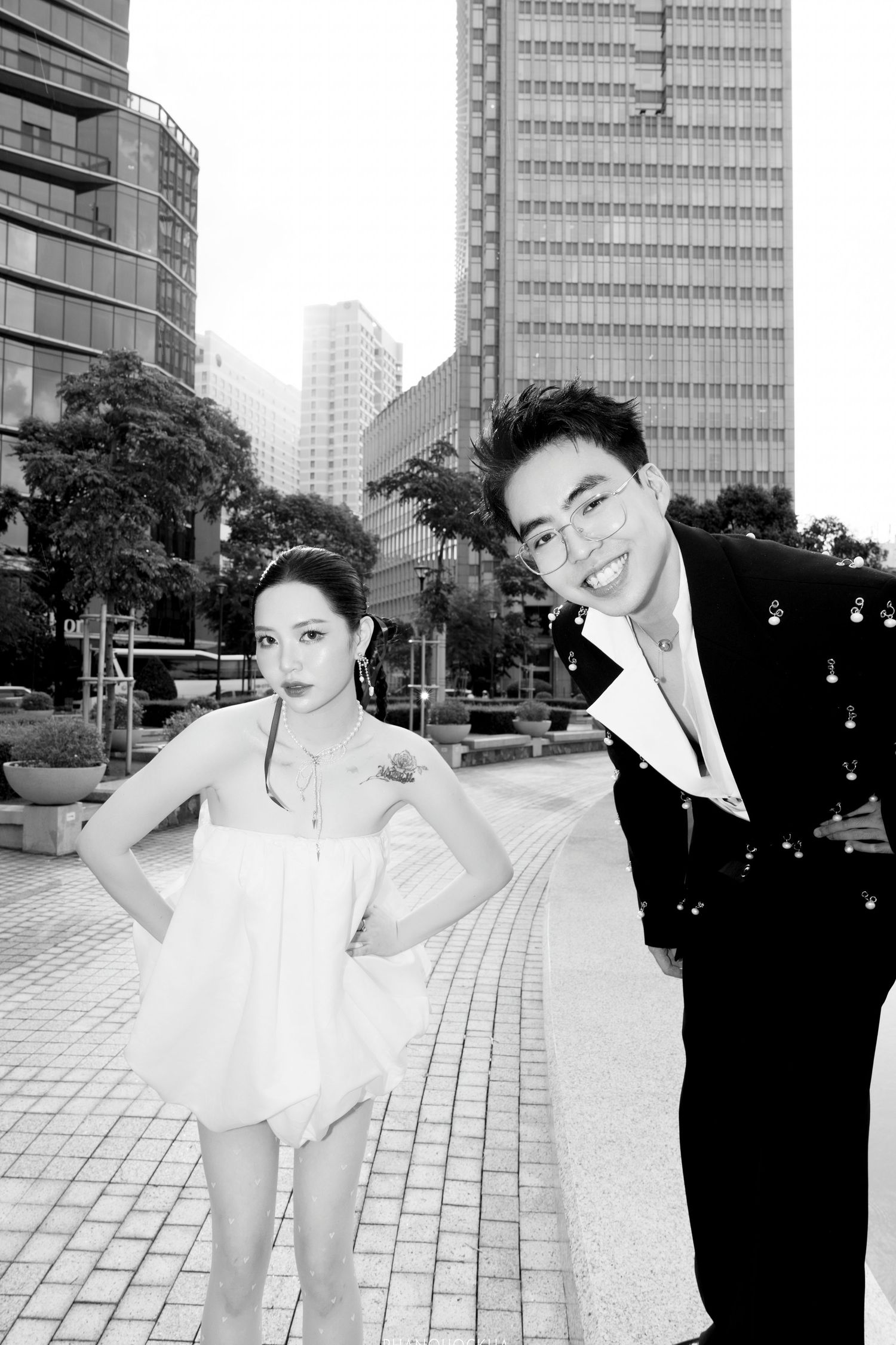 Black and white photo of stylish Asian couple posing together in front of city skyscrapers.