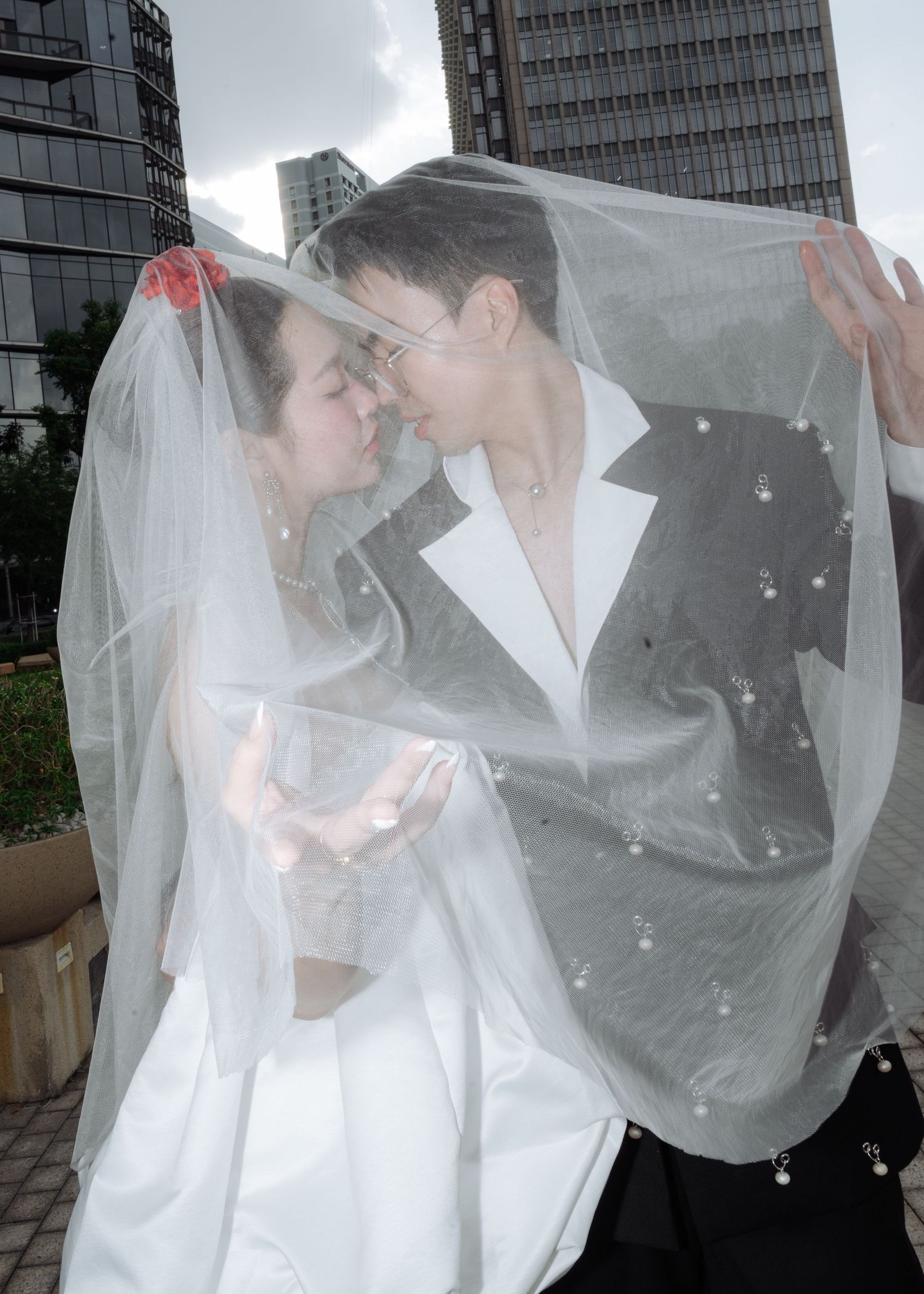 Bride and groom share a romantic moment under a pearl-adorned veil with city buildings behind them.